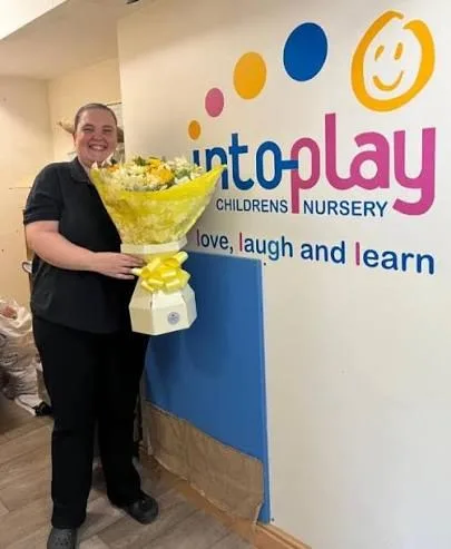 A member of the team holding flowers beside the Into-Play logo wall
